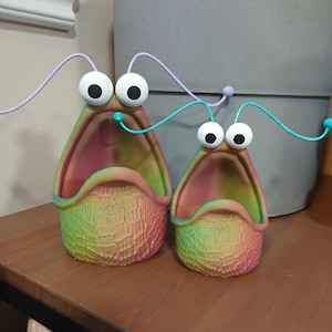 Könnte beinhalten: Zwei bunte Keramik-Pflanzer in Form von offenen Mündern mit Glitz Augen und Drahtantennen. Die Pflanzer sind grün, gelb und rosa.