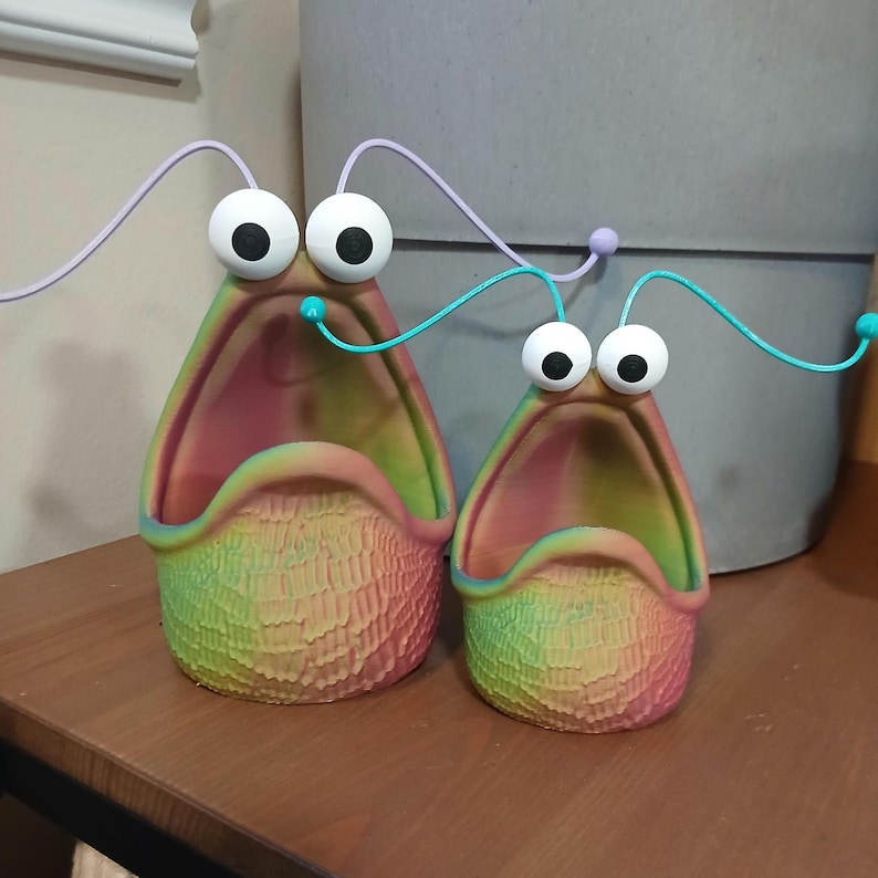 May include: Two colorful ceramic planters shaped like open mouths with googly eyes and wire antennae. The planters are green, yellow, and pink.
