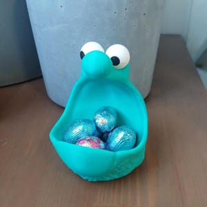 以下が含まれることがあります： 目は大きく口を開けたモンスターの形をしたターコイズブルーのセラミック製のイースターエッグホルダー。ホルダーはアルミホイルで包まれたチョコレートエッグでいっぱいです。