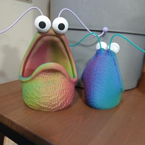 May include: Two colorful ceramic figurines with large eyes and open mouths. The larger figurine is rainbow colored and the smaller figurine is blue and green.