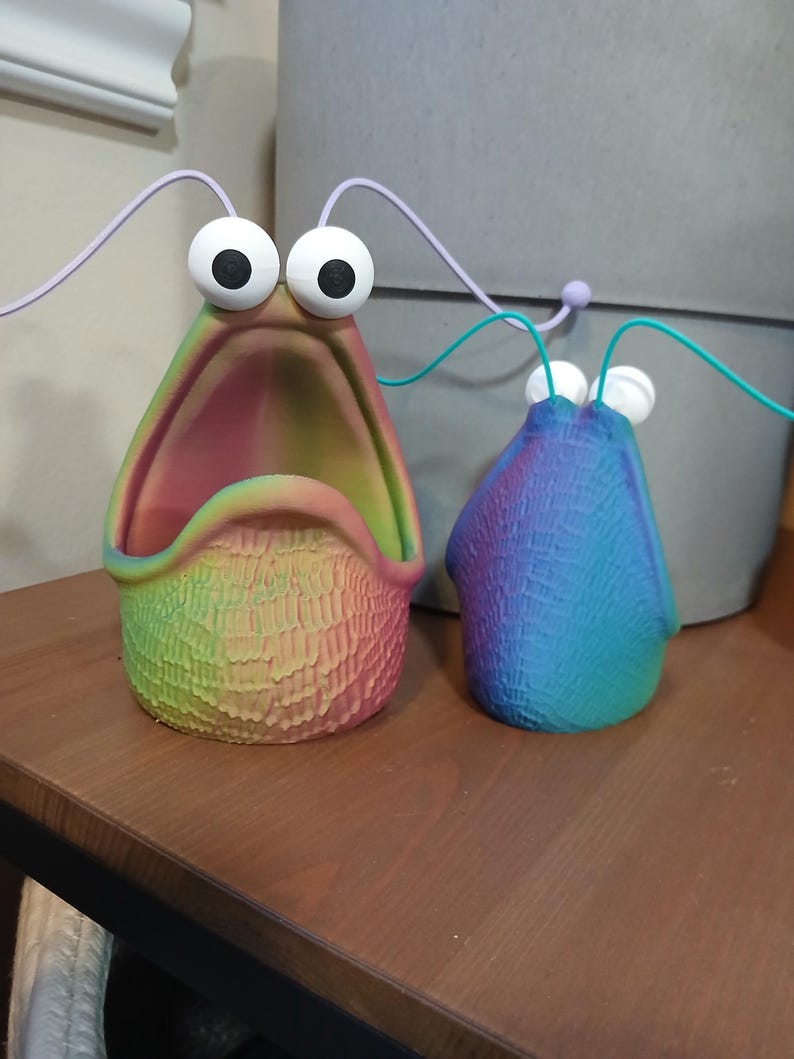 May include: Two colorful ceramic figurines with large eyes and open mouths. The larger figurine is rainbow colored and the smaller figurine is blue and green.