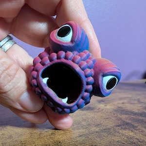 Puede incluir: Un objeto morado y azul con forma de criatura con múltiples ojos y una gran boca abierta. El objeto tiene una superficie texturizada y ojos grandes con bordes blancos y pupilas negras. El objeto se sostiene en una mano.