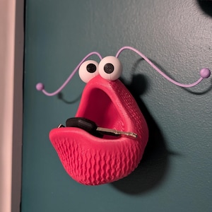 May include: A whimsical, bright pink key holder shaped like a mouth with large, googly eyes and antennae. The key holder is mounted on a teal wall and holds a black car key. The design is playful and unique.