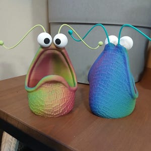 May include: Two colorful ceramic planters shaped like a creature with large eyes and an open mouth. The planters are decorated with a rainbow gradient and a textured surface.