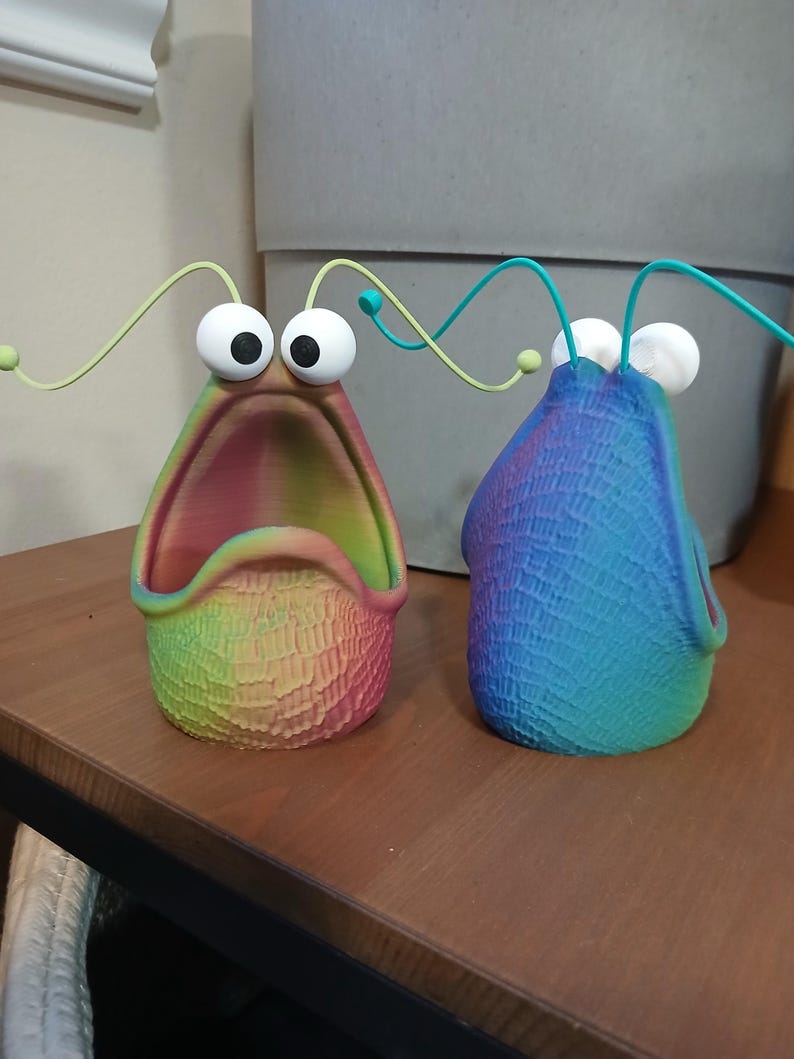 May include: Two colorful ceramic planters shaped like a creature with large eyes and an open mouth. The planters are decorated with a rainbow gradient and a textured surface.