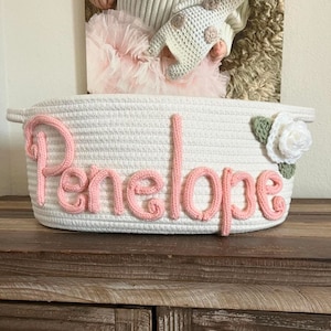May include: A white rope basket with the name "Penelope" in pink yarn lettering. The basket has a white rose and green leaves on the side. The basket is sitting on a wooden surface.