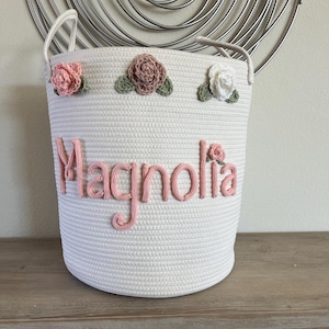 May include: White rope storage basket with pink and white crocheted flowers and the name "Magnolia" in pink yarn.
