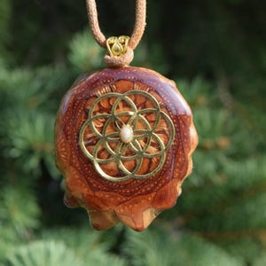 May include: A handcrafted pendant featuring a natural pinecone base with a gold-colored geometric design and a small, white bead at the center. The pendant hangs from a tan cord, set against a blurred green background.