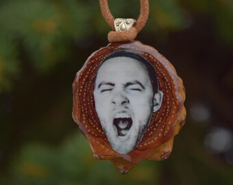 May include: A pendant featuring a black and white image of a person with their mouth open, set within a brown, organic-shaped frame. The pendant hangs from a brown cord with a silver-colored clasp.