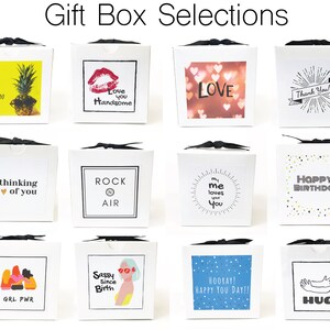 Peut inclure: Douze bo&icirc;tes cadeaux blanches avec diff&eacute;rents motifs et messages. Les bo&icirc;tes sont attach&eacute;es avec un ruban noir. Les motifs comprennent un ananas, une l&egrave;vre rouge, un c&oelig;ur, une &eacute;toile filante, un c&oelig;ur avec le mot "Love", un c&oelig;ur avec les mots "thinking of you", un motif noir et blanc avec les mots "Rock N Air", un soleil &eacute;clatant avec les mots "my me loves you", une &eacute;toile filante avec les mots "Happy Birthday", un motif avec les mots "GRL PWR", un motif avec les mots "Sassy Since Birth", un motif bleu avec les mots "Hooray! Happy You Day!!", et un motif avec les mots "Hug".