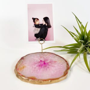 May include: A pink and gold agate photo holder with a gold wire clip, holding a photograph of a mother and child. The agate slice has a gold-painted edge and a vibrant pink center. A green plant is in the background.
