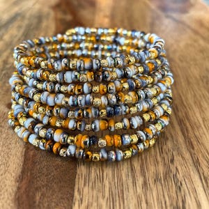Könnte beinhalten: Ein mehrreihiges Armband mit einer Mischung aus weißen, grauen, goldenen und orangefarbenen Perlen. Das Armband besteht aus kleinen, runden Perlen und ist mehrmals um sich selbst gewickelt.