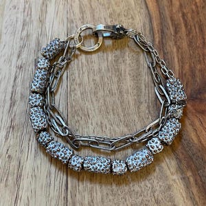 May include: A silver bracelet with two strands. One strand features textured, cylindrical beads with a speckled pattern. The other strand is a delicate chain. The bracelet has a clasp with the marking "925".