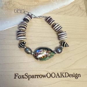 Op de afbeelding: Een armband met een centrale ovale iriserende abalone schelpkraal, geflankeerd door grijze parels, kleine gestreepte spiraalschelpen en platte ronde kralen met patronen in tinten paars, wit en bruin, afgewerkt met een zilverkleurige metalen ketting.