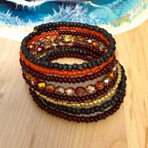 May include: A multi-strand wrap bracelet featuring a combination of matte black, glossy orange, deep burgundy, and metallic gold seed beads, interspersed with faceted amber-toned glass beads arranged in a circular, layered coil design.