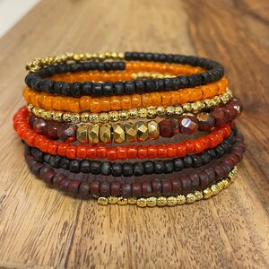 May include: A stack of colorful beaded bracelets in shades of orange, black, gold, and maroon. The bracelets are made of small, round beads and are arranged in a spiral pattern.