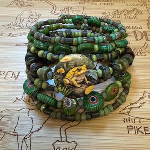 May include: A multi-strand coil bracelet featuring various shades of green beads in different shapes and textures. A central circular focal piece displays a detailed illustration of a yellow toad sitting among green foliage and yellow mushrooms.