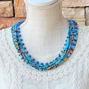 May include: A multi-strand beaded necklace in shades of blue, turquoise, orange, and brown. The necklace is displayed on a white mannequin. The beads are strung on a silver chain, creating a layered effect. The necklace is a statement piece.