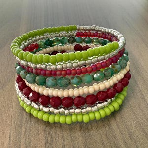 May include: A stack of beaded bracelets in various colors. The bracelets include green, red, silver, turquoise, and cream-colored beads. The beads are different shapes and sizes, creating a layered effect. The bracelets are stacked on top of each other.