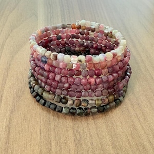 Op de afbeelding: Een stapel armbanden met kralen in tinten roze, rood en grijs. De armbanden zijn gemaakt van kleine, vierkant gesneden kralen, waardoor een textuur ontstaat. De kleuren variëren van lichtroze tot diep bordeauxrood en antracietgrijs.
