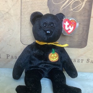 May include: Black plush beanie baby with a sparkly finish and a yellow ribbon around its neck. The bear has a black nose and black eyes. The bear is wearing a patch with a smiling orange pumpkin on it. The beanie baby has a Ty tag attached to its ear.
