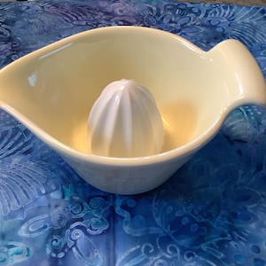 May include: Cream-colored ceramic citrus juicer with a built-in reamer. The juicer has a wide, curved bowl with a handle on one side and a spout on the other. The reamer is white and cone-shaped. The juicer sits on a blue patterned surface.