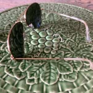 May include: Sunglasses with dark green lenses and gold-colored frames rest on a green ceramic plate with a grape and leaf design. The sunglasses have a classic aviator style.