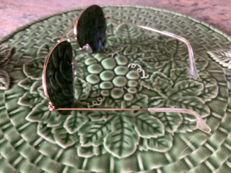May include: Sunglasses with dark green lenses and gold-colored frames rest on a green ceramic plate with a grape and leaf design. The sunglasses have a classic aviator style.