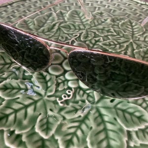 May include: Gold-framed aviator sunglasses with dark green lenses. The sunglasses have a classic design with a double bridge and a sleek, metallic finish. The lenses are reflective, and the frame is thin and lightweight.