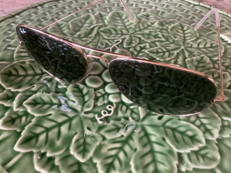 May include: Gold-framed aviator sunglasses with dark green lenses. The sunglasses have a classic design with a double bridge and a sleek, metallic finish. The lenses are reflective, and the frame is thin and lightweight.