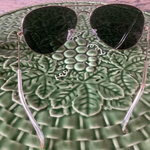 May include: Aviator sunglasses with a gold frame and dark green lenses. The sunglasses are resting on a green ceramic plate with a grape and leaf design. The plate has a woven basket pattern.