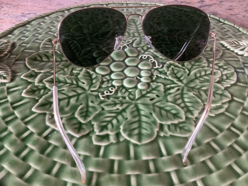 May include: Aviator sunglasses with a gold frame and dark green lenses. The sunglasses are resting on a green ceramic plate with a grape and leaf design. The plate has a woven basket pattern.