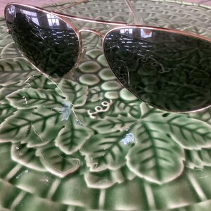 May include: Gold-framed aviator sunglasses with dark green lenses rest on a green ceramic plate with a leaf pattern. The sunglasses have a classic design, reflecting the surrounding environment.