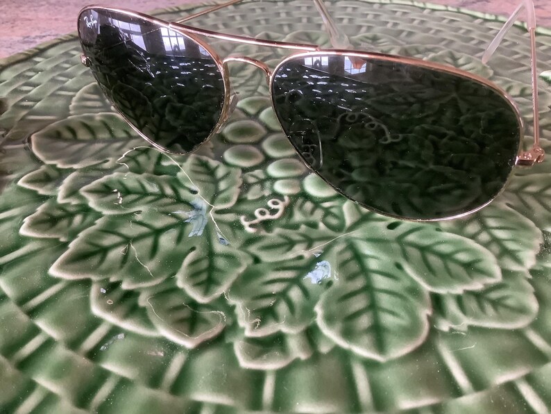 May include: Gold-framed aviator sunglasses with dark green lenses rest on a green ceramic plate with a leaf pattern. The sunglasses have a classic design, reflecting the surrounding environment.