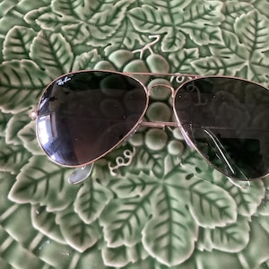 May include: Aviator-style sunglasses with a gold-colored frame and dark lenses. The Ray-Ban logo is visible on the upper left lens. The sunglasses are resting on a green, leaf-patterned surface.