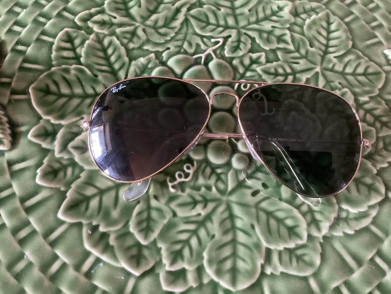 May include: Aviator-style sunglasses with a gold-colored frame and dark lenses. The Ray-Ban logo is visible on the upper left lens. The sunglasses are resting on a green, leaf-patterned surface.