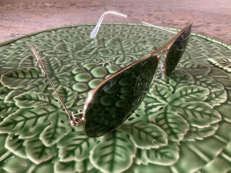 May include: Gold-framed sunglasses with green lenses, resting on a green, leaf-patterned surface. The sunglasses have a classic aviator shape and the Ray-Ban logo is visible on the lens.