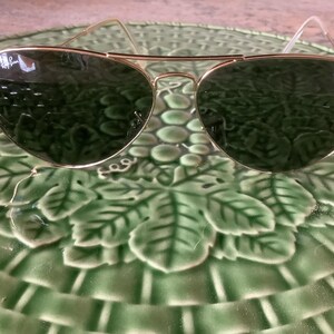 May include: Aviator-style sunglasses with gold-colored frames and dark green lenses. The sunglasses are resting on a green, leaf-patterned plate. The Ray-Ban logo is visible on the lens.