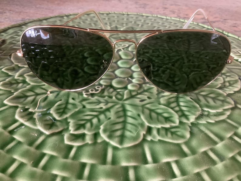 May include: Aviator-style sunglasses with gold-colored frames and dark green lenses. The sunglasses are resting on a green, leaf-patterned plate. The Ray-Ban logo is visible on the lens.