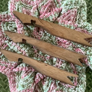 May include: Three wooden knitting needles with pointed ends, resting on a pink and green crocheted fabric.