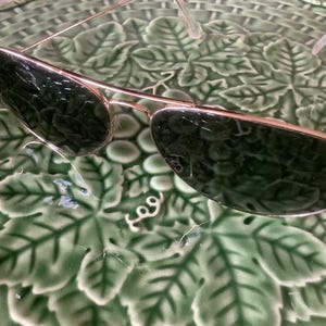 May include: Gold-framed sunglasses with dark green lenses. The sunglasses have a classic aviator shape and are resting on a green, leaf-patterned surface. The Ray-Ban logo is visible on the left lens.