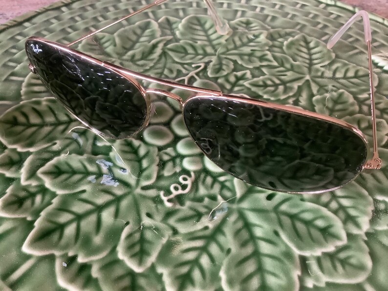 May include: Gold-framed sunglasses with dark green lenses. The sunglasses have a classic aviator shape and are resting on a green, leaf-patterned surface. The Ray-Ban logo is visible on the left lens.