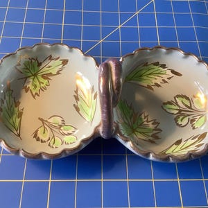 May include: A white ceramic double dish with a purple handle. The dish has a scalloped edge and is decorated with green and gold leaf designs.