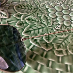 May include: Aviator sunglasses with a gold-colored frame and dark green lenses. The sunglasses are partially visible, resting on a green, grape-themed decorative plate. The lenses reflect the surrounding environment.