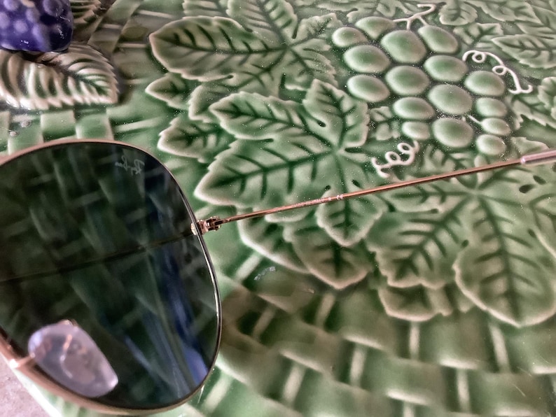 May include: Aviator sunglasses with a gold-colored frame and dark green lenses. The sunglasses are partially visible, resting on a green, grape-themed decorative plate. The lenses reflect the surrounding environment.