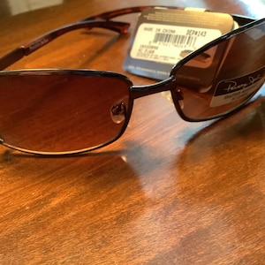 May include: Brown sunglasses with a dark frame and gradient lenses. The sunglasses have a decorative element on the temples and a tag attached. The brand name is visible on the tag.