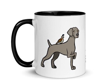 Könnte beinhalten: Eine weiße Keramik-Kaffeetasse mit schwarzem Rand und Henkel. Die Tasse zeigt eine Cartoon-Illustration eines Weimaraner-Hundes mit einem Wachtel, der auf seinem Rücken sitzt.