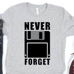 Puede incluir: Una camiseta gris con un gráfico de disquete negro y el texto "NEVER FORGET" impreso en ella.