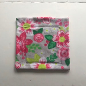 Puede incluir: Una almohadilla cuadrada de forro polar gris con un estampado floral. El diseño presenta flores rosas y blancas con hojas verdes y centros amarillos. La almohadilla tiene un borde cosido y un cuadrado empotrado en el centro.