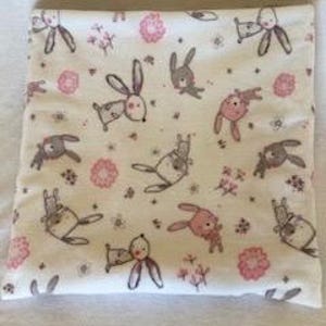 Puede incluir: Una manta de bebé blanca con un patrón de conejos rosas y grises. Los conejos sostienen flores y corazones.
